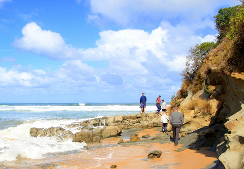 Beachgoers exploring rocky outcrops and sandy shore with eroded cliff face and ocean waves.