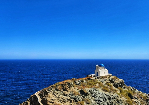 White chapel perched on a rocky outcrop overlooking a calm blue sea under clear sky.