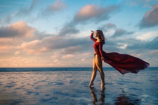 Woman in red dress standing in shallow ocean water with flowing fabric, hand raised to head, golden hour sky with clouds.