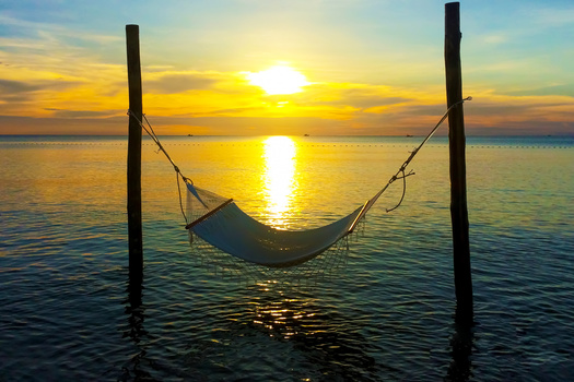 Empty hammock strung between two wooden posts suspended over calm ocean water at sunset.