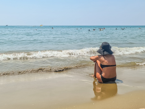 Woman in a blue hat and orange bikini sitting in shallow ocean water, looking toward the horizon.
