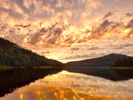 Lake at sunset with forested mountains reflected in still water under dramatic golden clouds.