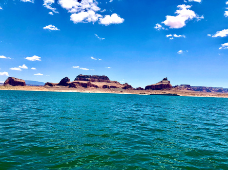 Turquoise water meets rust-colored desert cliffs under a bright blue sky with white clouds.