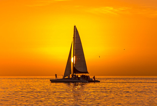 Silhouetted sailboat with full sail against a golden sunset over calm ocean waters.