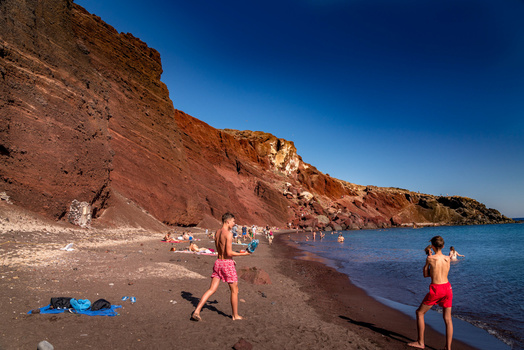 Beachgoers swimming and relaxing on a sandy beach beneath towering red and ochre cliffs under clear blue sky.