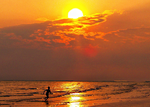 Silhouetted surfer wading into ocean at sunset with golden sun and dramatic clouds.