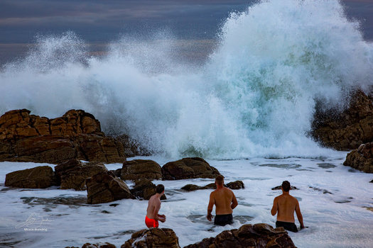 Three shirtless swimmers stand in shallow water as a massive wave crashes against rocky outcrops nearby.