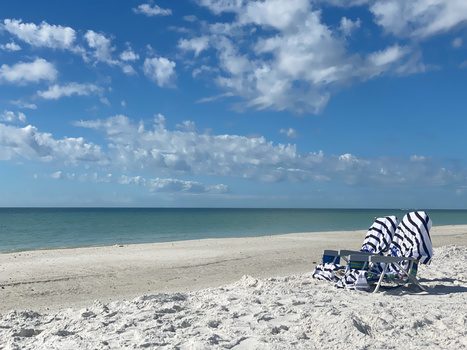 Beachgoer relaxing under a striped umbrella on a sandy shore with calm turquoise water and blue sky.