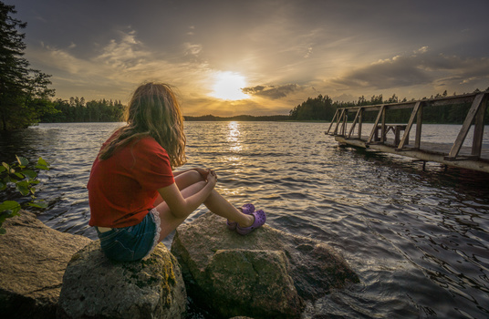 Woman in red shirt crouching on rocks at lakeside during golden hour sunset.