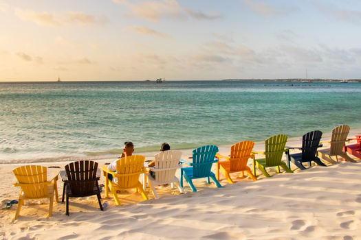 Colorful Adirondack chairs lined up on a sandy beach facing turquoise ocean waters.