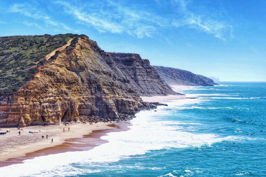 Dramatic coastal cliffs with golden-brown stratified rock formations rising above a white sand beach and turquoise ocean.