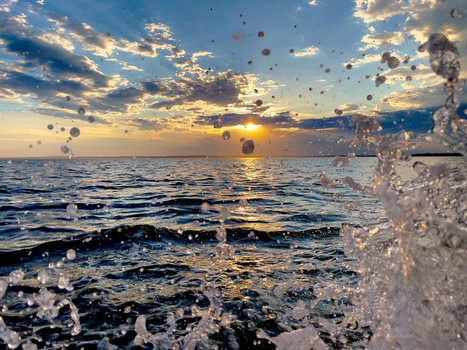 Ocean waves splashing in the foreground with sunset reflecting on water and clouds in the sky.