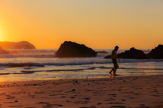 Silhouetted surfer carrying board on beach at golden hour sunset with rock formations in ocean.