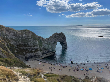 Natural rock arch formation jutting from a beach cove with tourists and blue water.