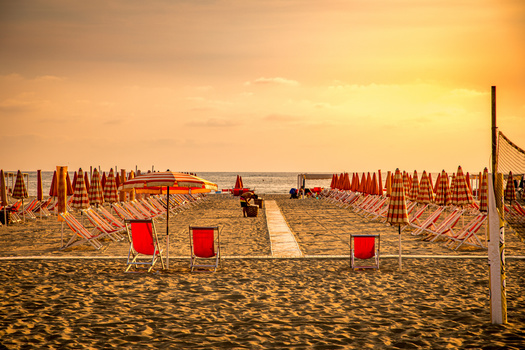Golden hour light illuminates an Italian beach lined with red umbrellas and wooden loungers stretching toward the horizon.