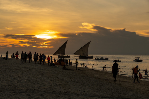 Silhouetted traditional sailing boats and beachgoers on shore during golden hour sunset.