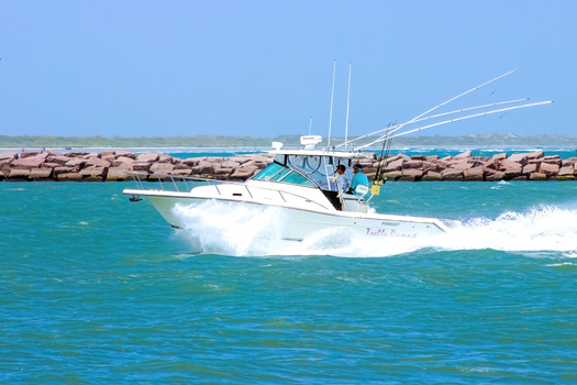 White fishing boat with outriggers cruising through turquoise water near a breakwater and pier.