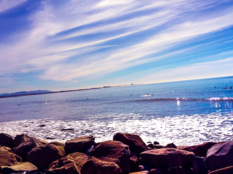 Rocky shoreline with waves breaking against dark boulders under a blue sky with wispy clouds.