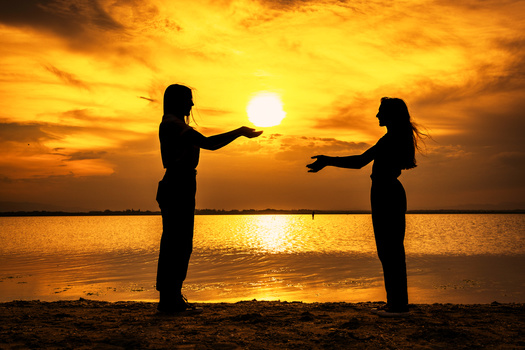 Two silhouetted figures reaching toward each other on a beach at golden hour sunset.