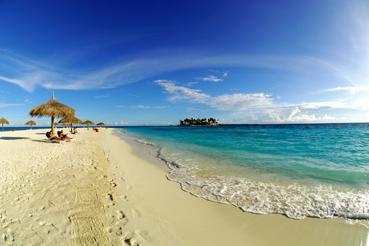 Tropical beach with white sand, turquoise water, and a thatched umbrella shelter on the left, with a small island visible on the horizon.