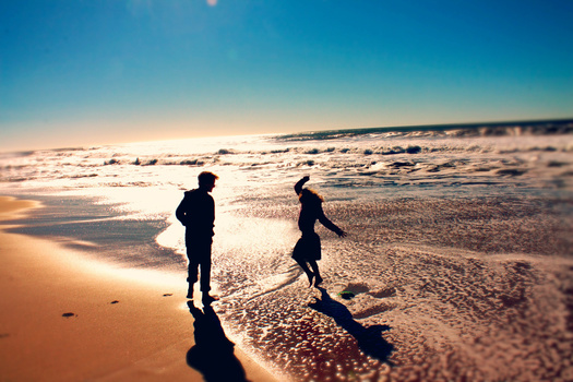 Two figures in dark clothing wading through shallow ocean waves at sunset or sunrise.