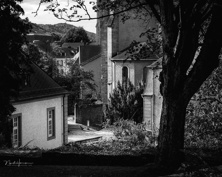Black and white photograph of white buildings nestled among trees, with a tall chimney rising between structures.