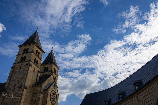 Twin-towered stone church with pointed spires against a bright blue sky with white clouds.
