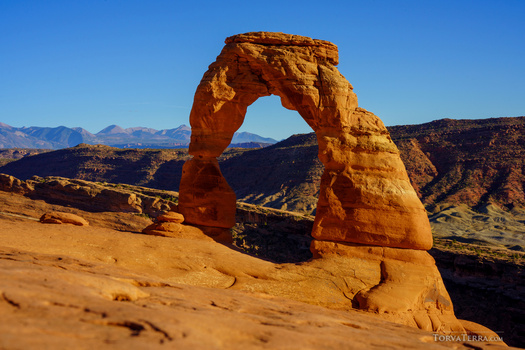 Natural stone arch formation framing distant snow-capped mountains under clear blue sky.