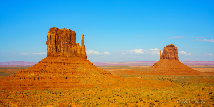 Two large red rock buttes rise from an expansive desert plain under a blue sky with scattered white clouds.