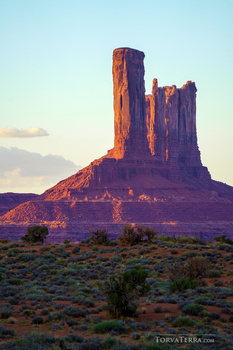 Tall red rock formation rising from desert landscape at golden hour with sparse vegetation in foreground.