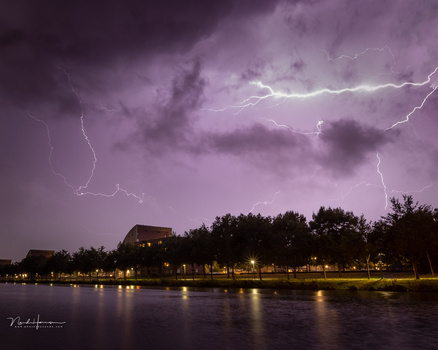 Lightning streaks across purple storm clouds above a riverside town at dusk, with buildings and trees illuminated below.
