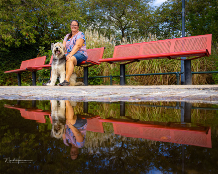 Person and large dog sitting on a red bench beside still water with trees reflected below.