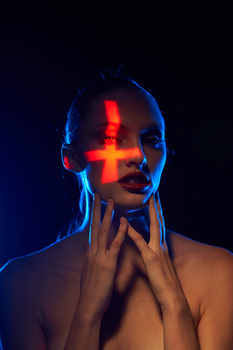 Woman's face illuminated by a glowing red cross projected onto her forehead and nose against dark blue background.