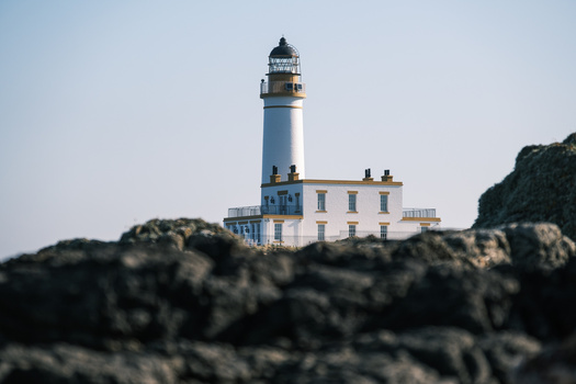 Blue and white lighthouse with attached keeper's house perched on rocky coastal outcropping.