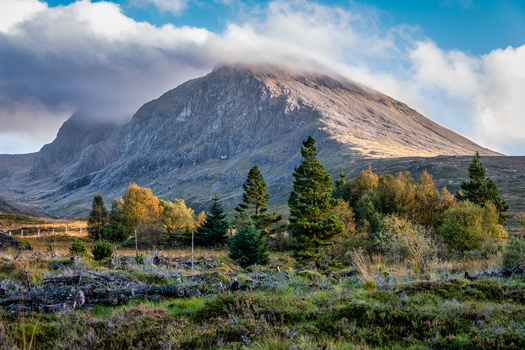 Conical mountain with rocky peak and forested slopes beneath partly cloudy sky.