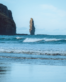 Tall sea stack rising from turquoise waters with breaking waves and dark cliffs framing the composition.