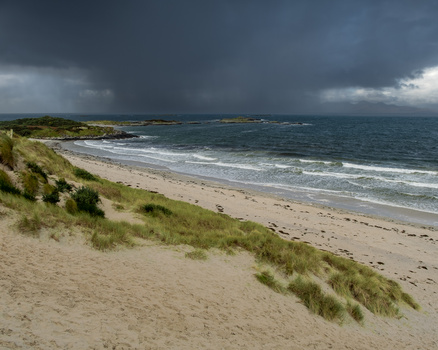Dramatic coastal landscape with sand dunes, beach, and stormy sky over rough ocean waters.