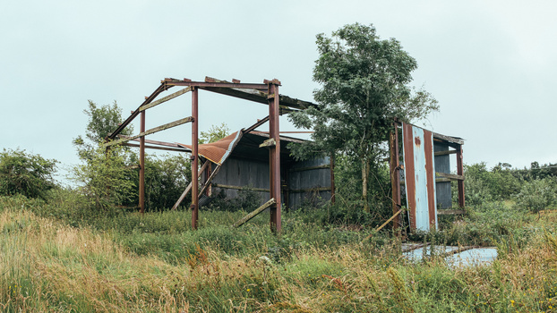 Abandoned agricultural structure with rusted metal framework overgrown by vegetation in a rural field.