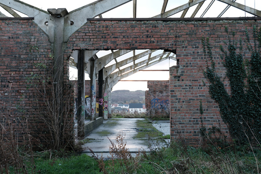 Symmetrical view through brick archway ruins with overgrown vegetation and industrial metal framework overhead.