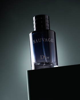 Dark fragrance bottle with black cap positioned on geometric black platform against teal gradient background.