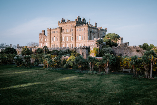 Red sandstone castle with multiple towers on a hilltop surrounded by green lawns and palm trees.