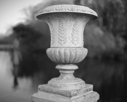Ornamental stone urn with decorative leaf relief and pedestal base in soft focus landscape setting.