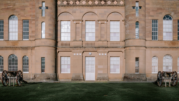 Symmetrical facade of a stone mansion with arched windows and decorative railings, flanked by two wooden wagons on grass.