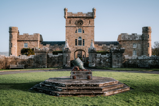 Historic stone castle with central clock tower and symmetrical wings, viewed across manicured lawn with cannon sculpture in foreground.