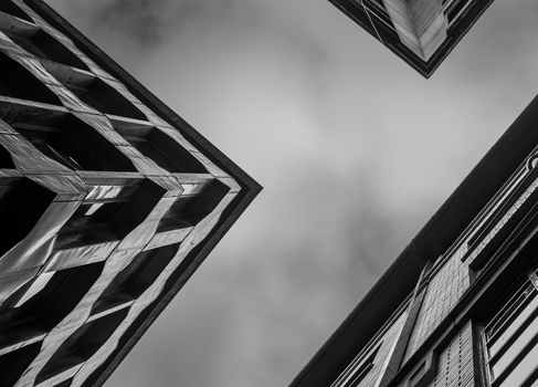 Black and white upward view of modern buildings with geometric facades against cloudy sky.