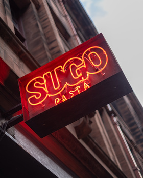 Red neon sign reading "STYGO PASTA" mounted on a building facade with vertical ribbed texture.