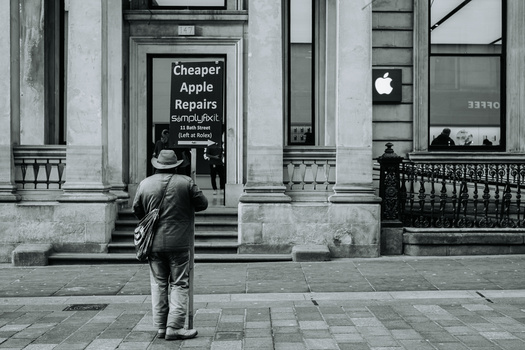 Man in hat and coat standing before a storefront with Apple Repairs sign on classical urban street.