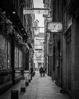 Narrow European street with historic storefronts and neon signs, rendered in high-contrast black and white.