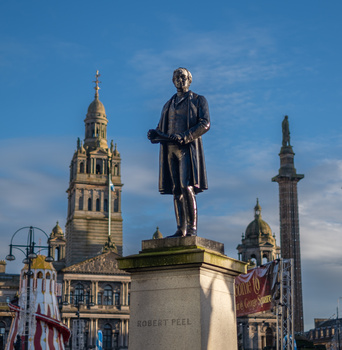 Bronze statue of a historical figure in period clothing stands centered against a clear blue sky, with ornate Victorian architecture and church spires in the background.