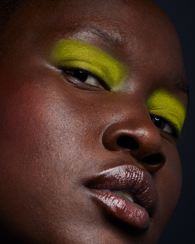 Close-up portrait of a person with vibrant lime-green eyeshadow and neutral lip color against dark background.